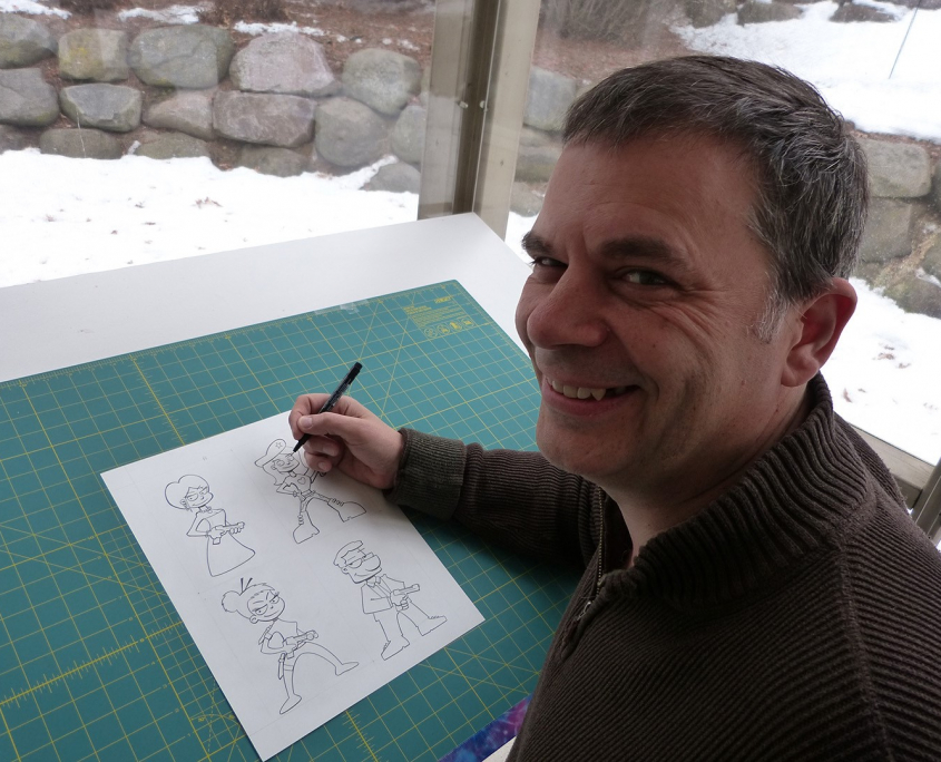 Photo of John Kovalic at drawing table with paper and cartoons he's drawn.