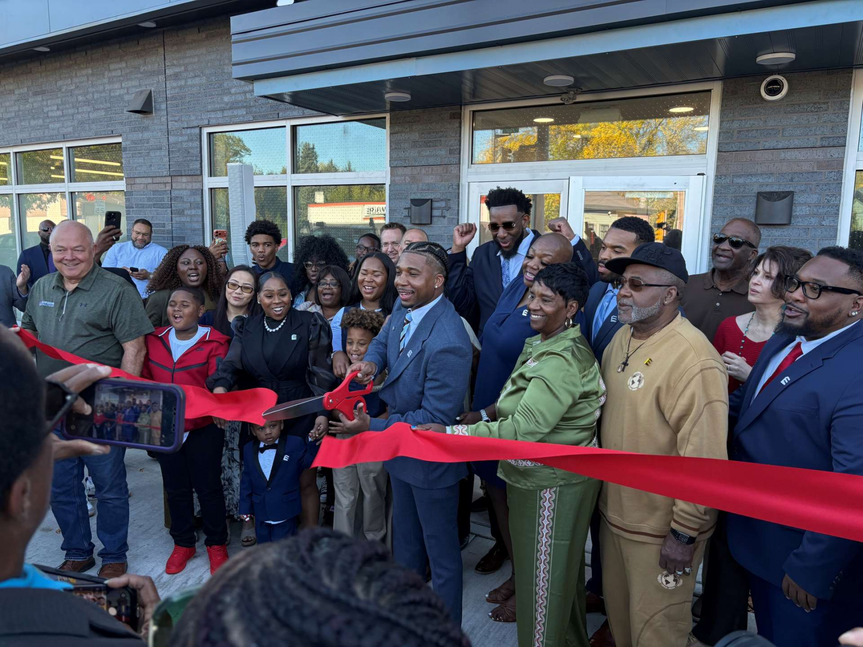 Man cutting ribbon in front of apartment building with a group of people around him.