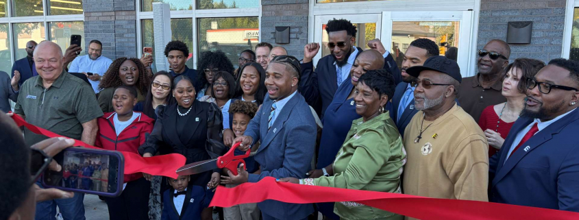 Man cutting ribbon in front of apartment building with a group of people around him.